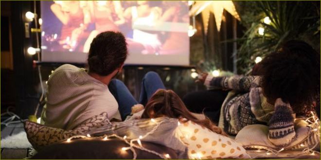 Společnost sledující film na promítacím plátně při večerním ležení venku ve světle loučí